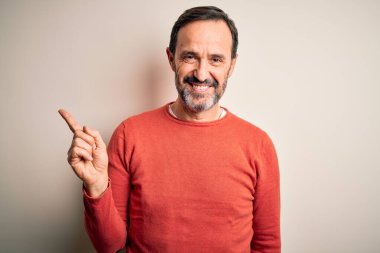 Orta yaşlı, turuncu bir süveter giyen, izole edilmiş beyaz arka planda duran, yüzünde kocaman bir gülümseme olan, el parmağı ile kameraya bakan, orta yaşlı bir adam..