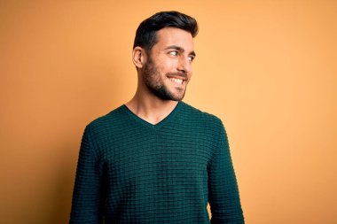 Sakallı, sıradan bir süveter giyen, sarı arka planda duran, yüzünde gülümseme, doğal bir ifade olan genç yakışıklı bir adam. Kendinden emin gülen.