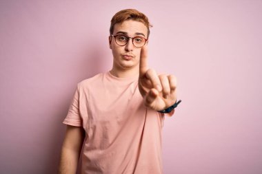 Genç, yakışıklı, günlük tişört giyen, izole edilmiş pembe arka plan üzerinde duran, parmağını kaldırmış, kızgın bir ifade sergileyen, hiçbir jest yapmayan bir adam.