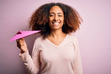 Afro saçlı, pembe arka planda elinde kağıt uçakla gülümseyen, kendine güvenen ve gülümseyen Afro-Amerikalı bir kadın.