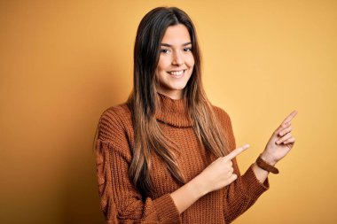 İzole edilmiş sarı arka planda dikilip gülümseyen ve kameraya bakan genç, güzel bir kız. İki eliyle ve parmaklarıyla işaret ediyor..