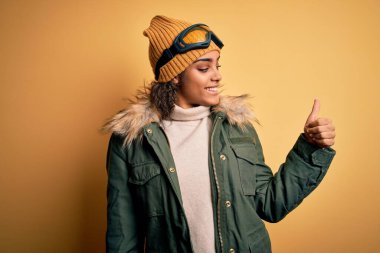 Genç Afrikalı Afro-Amerikan kayakçı kız kar giysileri ve kayak gözlüğü takıyor gururlu görünüyor, gülümsüyor yan tarafa başparmak hareketi yapıyor