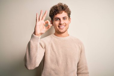 Sarışın, kıvırcık saçlı, beyaz arka planda rahat süveter giyen, gülümseyen, el ve parmaklarıyla imza atan genç, yakışıklı bir adam. Başarılı ifade.