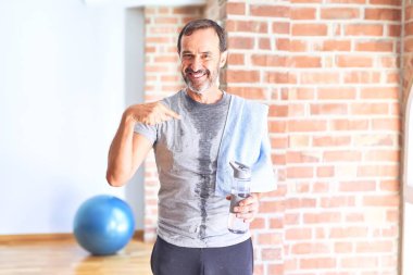 Orta yaşlı yakışıklı sporcu terli spor salonunda egzersiz yaptıktan sonra elinde bir şişe su ve havlu tutarken kendini gösteren sürpriz bir yüz ifadesiyle.