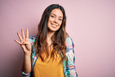 Genç, güzel, esmer, sıradan bir gömlek giyen, pembe arka planda duran, dört numarayı gösteren, kendinden emin ve mutlu bir şekilde gülümseyen bir kadın..