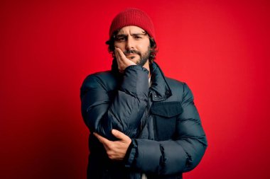 Sakallı, sıradan bir ceket giyen, kırmızı arka planda yün şapka takan genç ve yakışıklı bir adam kollarını kavuşturarak depresyondan sıkılmış ve yorgun görünüyor..