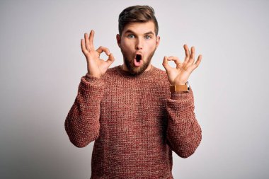 Sakallı, mavi gözlü, beyaz arka planda sıradan bir süveter giyen, şaşırmış ve şok olmuş genç bir adam parmaklarıyla onay sembolü yapıyor. Çılgın ifade