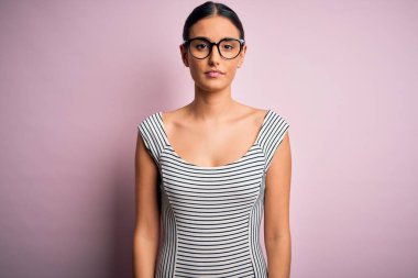 Genç ve güzel bir kadın pembe arka planda günlük çizgili elbise ve gözlük takıyor ve yüzünde ciddi bir ifade var. Basit ve doğal bir şekilde kameraya bakıyorsun..