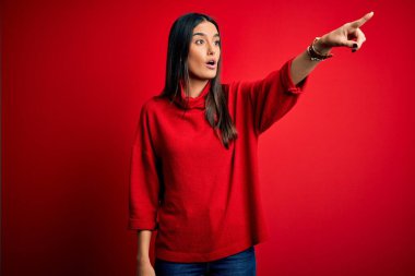 Genç, güzel esmer kadın izole edilmiş kırmızı arka plan üzerine gündelik süveter giyiyor. Parmağıyla işaret ediyor. İleride şaşırmış, ağzı açık, şaşırmış bir ifade.