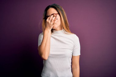 Mor arka planda sıradan tişörtler giyen, mavi gözlü genç, güzel, sarışın kadın yorgun, burnu ve gözleri ovuşturup yorgun ve baş ağrısı çekiyor. Stres ve hüsran kavramı.