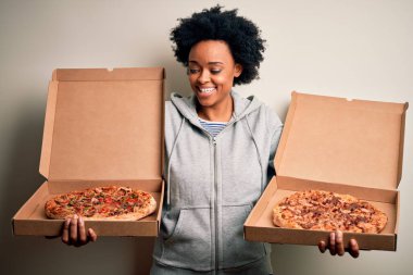Genç, güzel afro-amerikan afro kadın mutlu ve güvenle gülümsüyor. Yüzünde tebessümle durarak kutular dolusu İtalyan taze pizzasıyla izole beyaz arka planda.