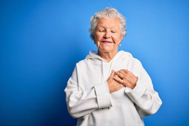 İzole edilmiş mavi arka plan üzerine beyaz kazak giyen, elleri göğsünde, gözleri kapalı ve yüzünde minnet dolu bir jest olan son sınıf güzeli bir kadın. Sağlık kavramı.