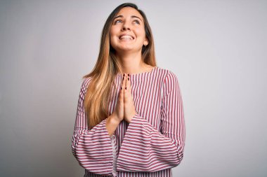 Mavi gözlü genç, güzel, sarışın bir kadın beyaz arka plan üzerinde ince tişört giyiyor, yalvarıyor ve dua ediyor. Yüzünde umut ifadesi var. Çok duygusal ve endişeli. Yalvarıyorum..