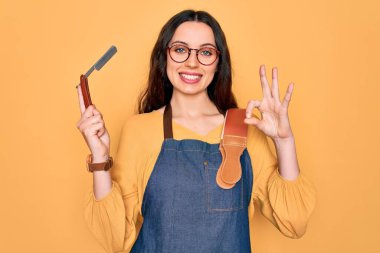 Önlük takan genç ve güzel berber kadın sarı arka planda jilet tutuyor. Tamam işareti yapıyor, mükemmel bir sembol.
