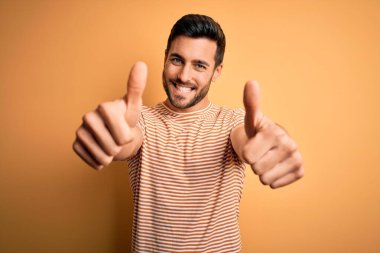 Sarı arka plan üzerine sıradan çizgili tişört giyen sakallı genç yakışıklı adam el hareketleriyle olumlu jest yapmayı onaylıyor, gülümsüyor ve başarı için mutlu. Kazanan jest.