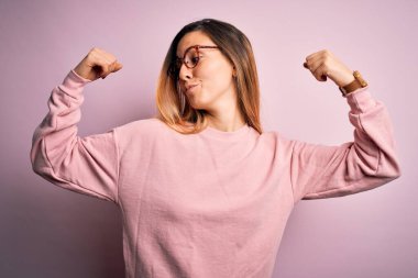 Mavi gözlü, pembe arka planda süveter ve gözlük takan güzel sarışın kadın, gururla gülümseyen kol kaslarını gösteriyor. Fitness konsepti.