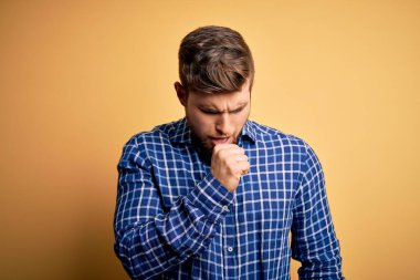 Sarışın, sakallı ve mavi gözlü genç bir işadamı sarı arka planda gömlek giyiyor ve soğuk algınlığı veya bronşitin belirtisi olarak öksürüyor. Sağlık hizmeti kavramı.