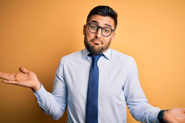 Sarı arka plan üzerinde duran kravat ve gözlük takan genç yakışıklı işadamı elleri ve kolları havada, kafası karışmış bir ifade takınıyor. Şüphe kavramı.