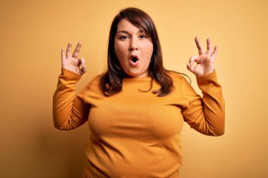 Güzel esmer ve cüsseli bir kadın izole edilmiş sarı arka plan üzerine günlük süveter giyiyor. Şaşırmış ve şaşırmış görünüyor. Tamam sembolünü parmaklarıyla yapıyor. Çılgın ifade