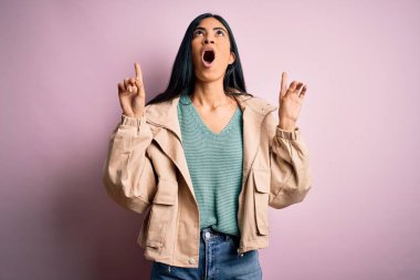 Pembe arka plan üzerinde kışlık ceket ve süveter giyen genç, güzel İspanyol moda kadını hayrete düşmüş ve şaşırmış bir şekilde parmaklarıyla işaret ediyor ve kollarını kaldırıyordu..