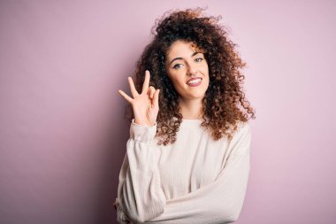 Kıvırcık saçlı ve piercingli güzel bir kadın pembe arka planda rahat bir süveter giyiyor ve üç numaralı parmaklarını gösterip gülümserken mutlu ve kendinden emin..