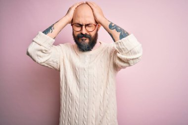 Sakallı, dövmeli, gözlüklü, pembe arka planda kazak giyen, başı ağrılı, çaresiz ve stresli yakışıklı bir adam. Çünkü ağrı ve migren. Ellerini başının üstüne koy..