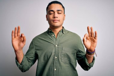 İzole edilmiş beyaz arka plan üzerinde dikilen Brezilyalı genç adam rahatla ve parmaklarıyla meditasyon hareketi yaparken gözleri kapalı gülümse. Yoga konsepti.