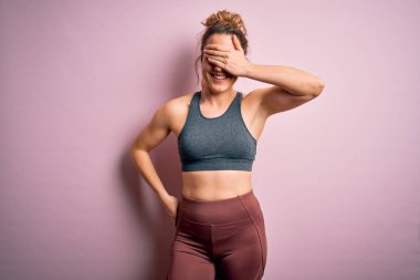 Genç ve güzel sarışın sporcu kadın spor yapıyor. Pembe arka planda spor kıyafetleri giyiyor. Gülümsüyor ve yüzünde şaşkınlıktan gözleri kapayarak gülüyor. Kör kavram.