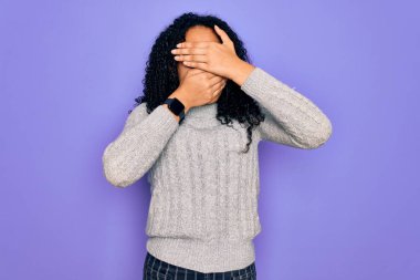 Genç Afro-Amerikalı kadın mor arka plan üzerine günlük süveter ve gözlük takıyor gözleri ve ağzı kapalı, şaşırmış ve şok olmuş. Duygularını gizliyor.