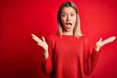 Kırmızı izole edilmiş arka plan üzerine gündelik süveter giyen genç, güzel sarışın kadın çılgın ve başarıyı kollarını kaldırıp heyecanla haykırarak kutluyor. Kazanan konsept