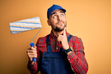 Genç yakışıklı ressam duvarları boyuyor. İzole edilmiş sarı arka plan üzerinde silindir kullanıyor. Ciddi bir yüz.