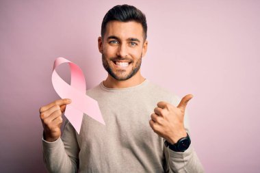Arkaplanda pembe kanser kurdelesi taşıyan yakışıklı bir adam. Büyük bir gülümsemeyle mutlu. İşaret ver, parmaklarınla başparmağını kaldır.