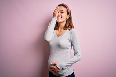 Genç, güzel bir kız hamile, pembe arka plan üzerinde bebek bekliyor. Bir gözünü elinle kapatıyor. Yüzünde kendine güvenen bir gülümseme ve sürpriz bir duygu var..