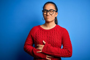 Genç, güzel Asyalı bir kadın mavi arka planda günlük süveter ve gözlük takıyor. Midesinde hazımsızlık, acı verici bir hastalık var. Ache kavramı.