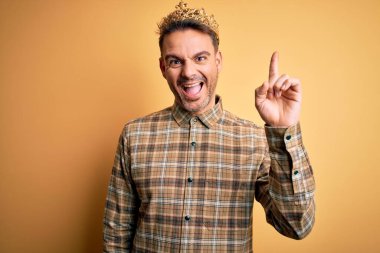 Sarı arka plan üzerinde kralın altın tacını takan yakışıklı genç adam başarılı bir fikirle işaret ediyor. Heyecanlı ve mutlu. Bir numara..