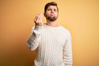 Sarışın, sakallı, mavi gözlü, sarı arka planda beyaz süveter giyen, el ve parmaklarıyla İtalyan jestleri yapan genç bir adam.