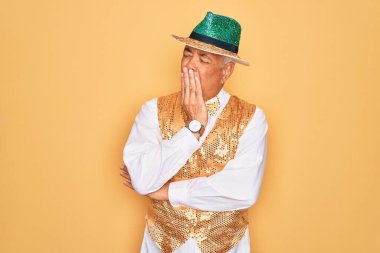 Sarı arka planda Brezilya karnavalı kıyafeti giyen orta yaşlı, kır saçlı bir adam esnemekten sıkılmış, ağzı eliyle kapatmaktan sıkılmış. Huzursuz ve uykulu..