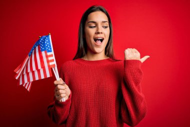 Amerika Birleşik Devletleri bayrağını kırmızı arka planda tutan güzel, vatansever bir kadın gülümseyen bir gülümsemeyle baş parmağıyla işaret ediyor.