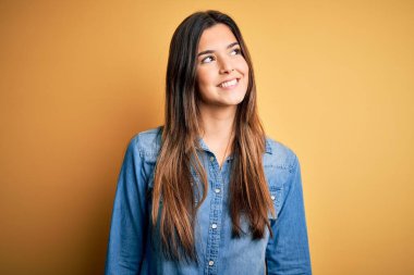 Genç, güzel bir kız, sıradan bir kot gömlek giyer, izole edilmiş sarı arka plan üzerinde durur, yüzünde gülümseme, doğal bir ifade ile yan yana bakar. Kendinden emin gülen.