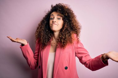 Kıvırcık saçlı, piercingli, zarif ceketli, kafası karışık, kolları ve elleri havada genç bir iş kadını. Şüphe kavramı.