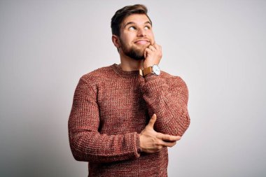 Sakallı ve mavi gözlü genç bir adam beyaz arka planda sıradan bir süveter giyiyor ve elleri çenesinde soru sormayı düşünüyor, düşünceli bir ifade. Gülen ve düşünceli bir yüz. Şüphe kavramı.