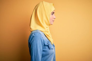 İslami tesettür giyen genç ve güzel bir kız yalıtılmış sarı arka planda duruyor, yan yana bakıyor, rahat bir görünüm sergiliyor, kendine güvenen bir gülümsemeyle doğal bir yüz ifadesi sergiliyor..