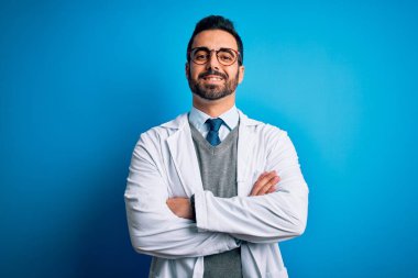 Sakallı, mavi arka planda gözlüklü, mutlu yüzlü, kollarını kavuşturup kameraya bakan yakışıklı bir doktor. Pozitif kişi.