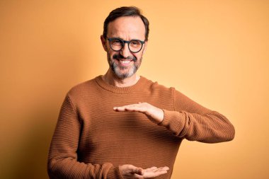 Orta yaşlı, beyaz, kahverengi süveter giyen ve gözlüklü, izole edilmiş sarı arka planda, elleri büyük ve büyük boy işareti olan, ölçü sembolü gösteren bir adam. Kameraya bakıp gülümsüyor. Ölçüm konsepti.