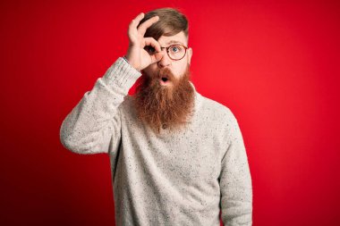Sakallı, kırmızı arka planda günlük süveter ve gözlük takan yakışıklı kızıl saçlı adam şaşırmış yüz ifadesiyle şaşırmış, parmaklarından bakan gözler. İnançsız ifade.