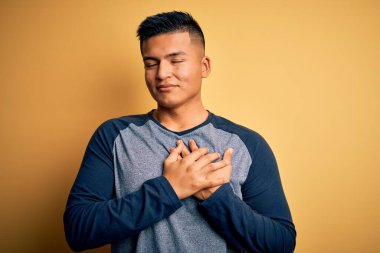 Sarı arka planda sıradan tişörtler giyen genç, yakışıklı Latin adam elleri göğsünde, gözleri kapalı ve yüzünde minnet dolu bir jest ile gülümsüyor. Sağlık kavramı.