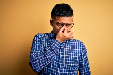 Sarı arka plan üzerine gündelik tişört ve gözlük takan genç, yakışıklı Latin adam yorgun burnu ve gözleri ovuşturarak yorgunluk ve baş ağrısı hissediyor. Stres ve hüsran kavramı.