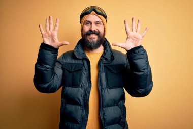 Sakallı, kar maskesi takmış, kar maskesi takmış, kel yakışıklı kayakçı on numaralı parmaklarıyla onu gösterirken kendinden emin ve mutlu bir şekilde gülümsüyor..