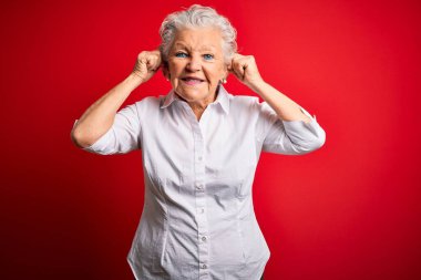 Son sınıf güzeli, zarif bir gömlek giymiş kırmızı arka plan üzerinde durmuş parmaklarıyla kulaklarını çekerek gülümseyen, komik hareketler yapan bir kadın. Seçme sorunu