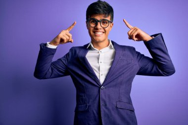 Ceket ve gözlük takan yakışıklı iş adamı mor arka planda gülümsüyor iki eliyle başını işaret ediyor, harika bir fikir ya da düşünce, iyi bir hafıza.